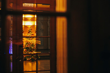 Creative view on stylish christmas tree with garland lights and  christmas decorations through window in european city street. Festive decorations and illumination in winter holidays in town