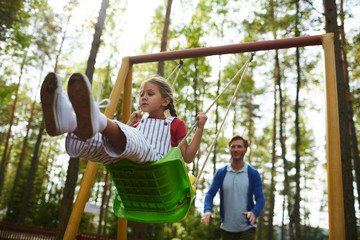 Wall Mural - Little girl sitting on swing while her father pushing her from behind during chill in park