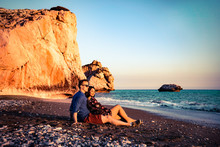 Young Loving Couple Sitting Together At The Beach In Front Of The Aphrodite Rock In Cyprus Greece During Sunset.