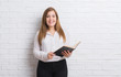 © Krakenimages.com - Young adult woman standing over white brick wall reading a book with a happy face standing and smiling with a confident smile showing teeth