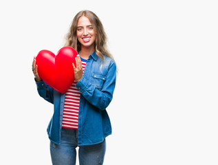 Wall Mural - Beautiful young blonde woman holding heart valentine over isolated background with a happy face standing and smiling with a confident smile showing teeth