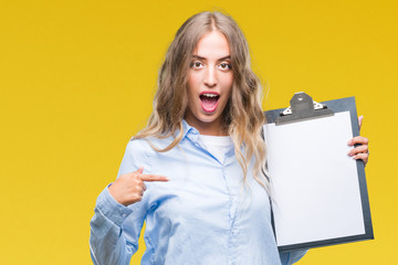 Wall Mural - Beautiful young blonde business woman holding clipboard over isolated background with surprise face pointing finger to himself