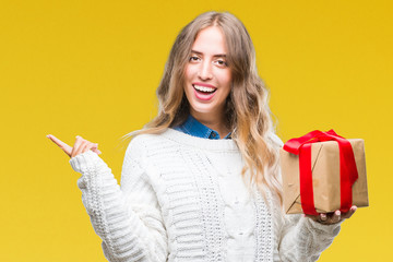 Canvas Print - Beautiful young blonde woman holding gift over isolated background very happy pointing with hand and finger to the side