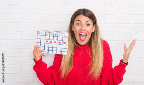 Fotografía Young adult woman over white brick wall holding period calendar very happy and e