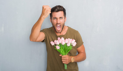 Handsome young man over grey grunge wall holding flowers bouquet annoyed and frustrated shouting with anger, crazy and yelling with raised hand, anger concept