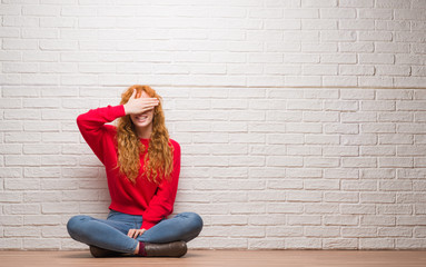 Canvas Print - Young redhead woman sitting over brick wall smiling and laughing with hand on face covering eyes for surprise. Blind concept.