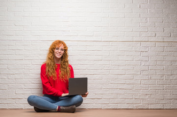 Poster - Young redhead woman sitting over brick wall using computer laptop with a happy face standing and smiling with a confident smile showing teeth