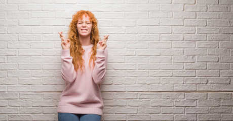 Wall Mural - Young redhead woman standing over brick wall smiling crossing fingers with hope and eyes closed. Luck and superstitious concept.