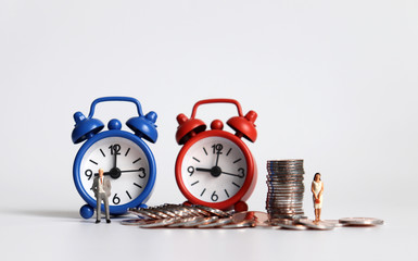 A miniature man standing on the blue alarm clock and a miniature woman standing on the red alarm clock with pile of coins.