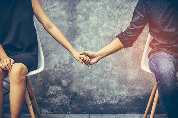 Young couples sitting on a chair inside the room which walls are made from cement. They shake hands, show their love and encouragement toward each other with copy space.