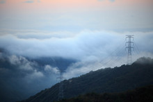 Towers Carrying High Voltage Electrical Lines Through Mountains And Fog At Sunrise