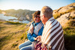 © Suzi Media  - Loving mature couple hiking, sitting on windy top of rock, exploring. Active Mature man and woman wrapped in blanket, hugging and Happily smiling. Scenic view of sea, mountains. Norway, Lindesnes.