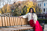 Fototapeta Krajobraz - Attractive brunette woman in red dress sitting on bench in autumn park