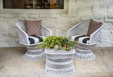 White Wicker Chairs And Table Outside On Porch With Autumn Flower Arrangement With Deer Horns All Against White Painted Stone Wall With Table And Candles Visable Through Window