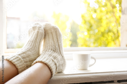 Fotomural Young woman sitting with crossed legs in warm woolen socks leaning on windowsill, cup of coffee and book, autumnal window view w/ yellow leaves