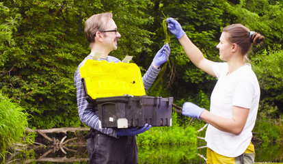 Man and woman scientist environmentalist standing in a river. Woman taking sample of duckweed. Man holding toolbox