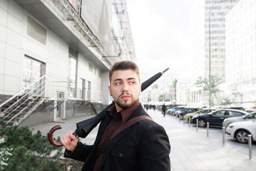 Portrait of a man with a beard and umbrella in the hands of a street background and modern architecture. Business man walks in the rain in the city and looks to the side.