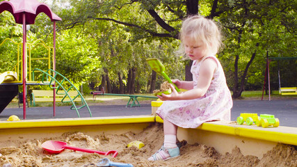 Low angle view. A little girl sitting in a sandbox is picking up sand with a scoop in a mold