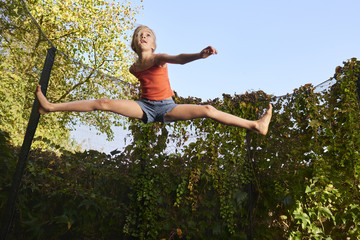 Wall Mural - Child cute blond girl playing and jumping on trampoline with greenery background