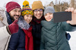 © Seventyfour - Waist up portrait of four happy young people taking selfie photo and showing thumbs up while having fun outdoors in winter