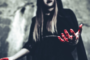 Fototapeta witch showing bloody hand. female demon angel in black clothes and hood on grunge wall background. halloween day and mystery concept. fantasy of magic theme. afterlife and death concept.