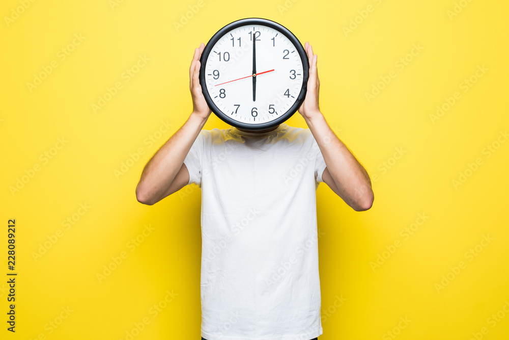 Young man holding big clock covering his face on yellow background - obrazy, fototapety, plakaty 