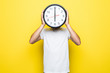 Young man holding big clock covering his face on yellow background