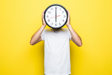 Young man holding big clock covering his face on yellow background