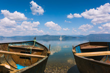 Two Old Fishing Boats And A Sailing Ship In The Distance. Blue Lake With Stones Near The Shore And Clouds Reflecting In The Water. Mountains In The Distance, Also Reflecting In The Water. Sunny Day.