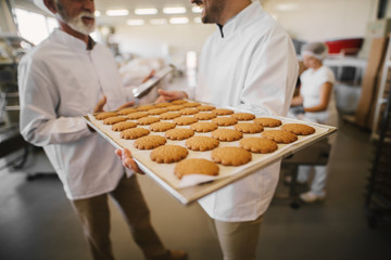 close up of fresh cookies on big tray in food factory. blurred picture of two male employees in ster