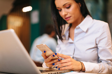 Young woman using smartphone in a cafe with a laptop in front of her