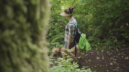 young woman and dog walking down a hiking trail in a forest with a mossy tree in the foreground.
