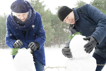 Portrait Of Modern Senior Couple Building Snowman While Enjoying Walk In Beautiful Winter Forest With Snow Falling Gently