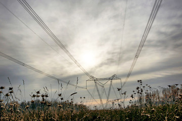 Autumn landscape with tall metal towers of high voltage electrical line against the cloudy sky and with dry grass on foreground