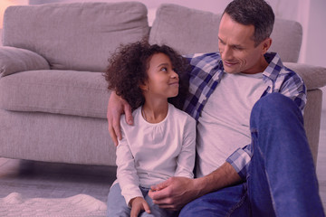 Wall Mural - Happy fatherhood. Positive nice man hugging his daughter while sitting with her in front of the sofa