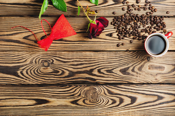 Full cup of black coffee near one red rose and scattered coffee beans and gift on old wooden table. Top view with copy space. Saint Valentine's Day concept