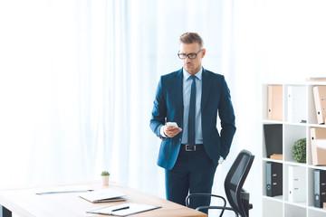 Wall Mural - handsome businessman in eyeglasses and formal wear using smartphone at workplace