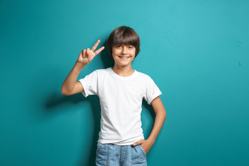 Smiling little boy in t-shirt showing Victory gesture on color background