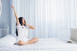 © anon - Woman stretching in bed after waking up, back view. Woman sitting near the big white window while stretching on bed after waking up with sunrise at morning, back view.