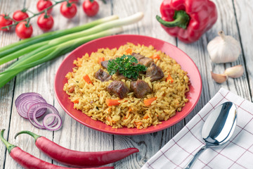 Wall Mural - Red plate with pilaf on a white wooden table. On the table is red pepper, green onions, garlic, cherry tomatoes, white napkin, spoon.