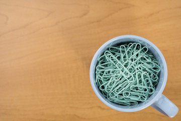 PASTEL GREEN PAPER CLIPS IN BLUE COFFEE CUP ON WOODEN OFFICE TABLE WITH COPY SPACE