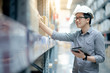 © zephyr_p - Young Asian man worker wearing safety helmet and eyeglasses doing stocktaking of product in cardboard box on shelves in warehouse by using digital tablet and pen. Physical inventory count concept