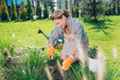 © zinkevych - Grubbing weeds up. Appealing family woman wearing squared shirt and orange gloves sitting on her knees while grubbing up weeds