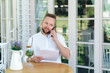 © romannoru - Close-up of a business man sitting at a table in a cafe, holding some important documents in his hand, talking on the phone with his personal assistant. The businessman has just signed an important