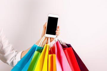 Cropped shot of female hand holding bunch of different colorful blank shopping bags and blank screen mobile phone over isolated white background. Black friday sale concept. Copy space, close up.