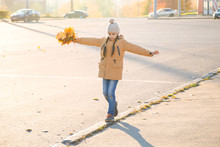 Positive Little Girl Walking On The Curb With Maple Leaves In Her Hand And Trying To Keep Her Balance. Autumn Cityscape