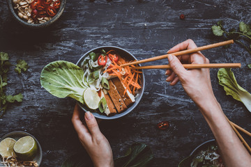 Eating delicious asian bowl with rice noodles, vegetables and tofu on wooden background. Top view