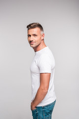 Wall Mural - side view of handsome man in white t-shirt smiling at camera isolated on grey