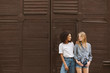 © Anton - Young nice african american woman with dark curly hair in T-shirt and jeans and pretty woman with blond hair in shirt and shorts happily looking at each other with brown wall on background