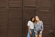 © Anton - Young beautiful african american woman with dark curly hair in T-shirt and jeans and woman with blond hair in shirt and denim shorts dreamily looking in camera with brown wall on background